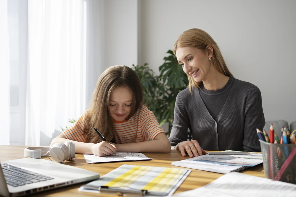 young girl writing on a paper at a desk with a tutor or mother watching and smiling