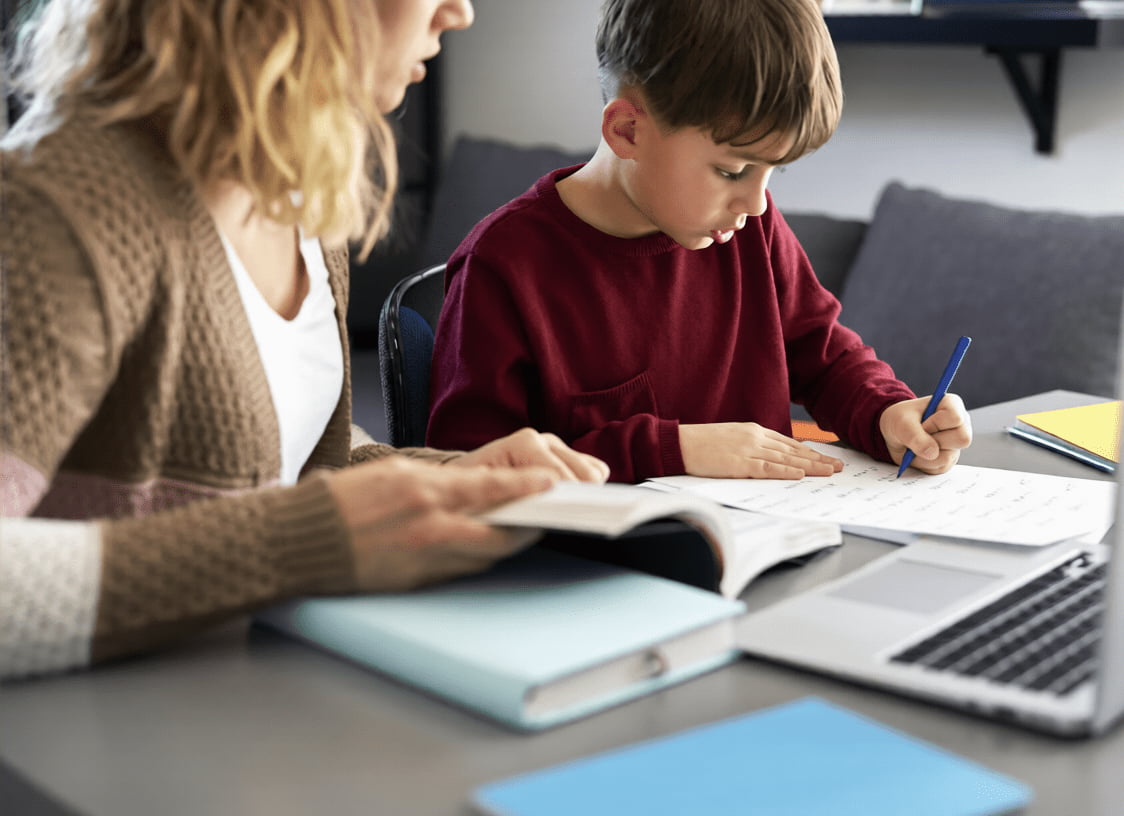young boy doing schoolwork at a desk with a tutor or parent guiding him